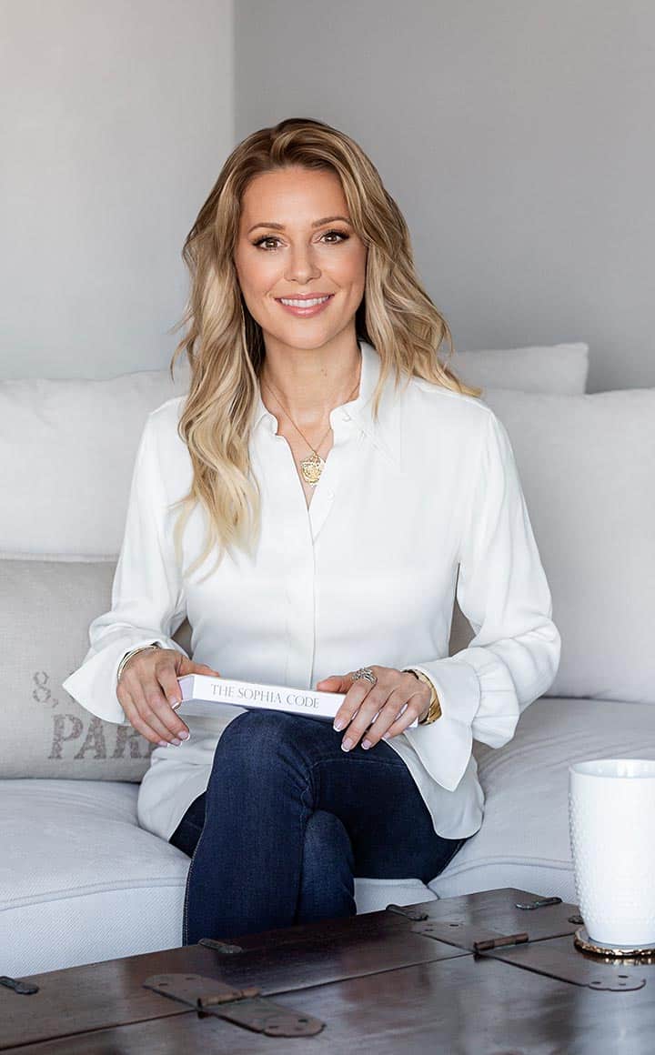 Kaia Ra in white shirt and black pants with gold pendant necklace sitting on white sofa with a copy of The Sophia Code on her lap