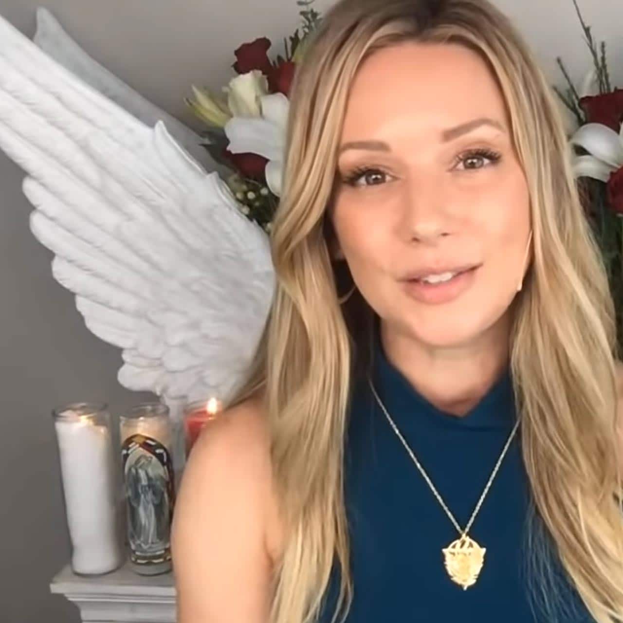 Kaia Ra in blue top with gold necklace in front of candles, wings, and flowers